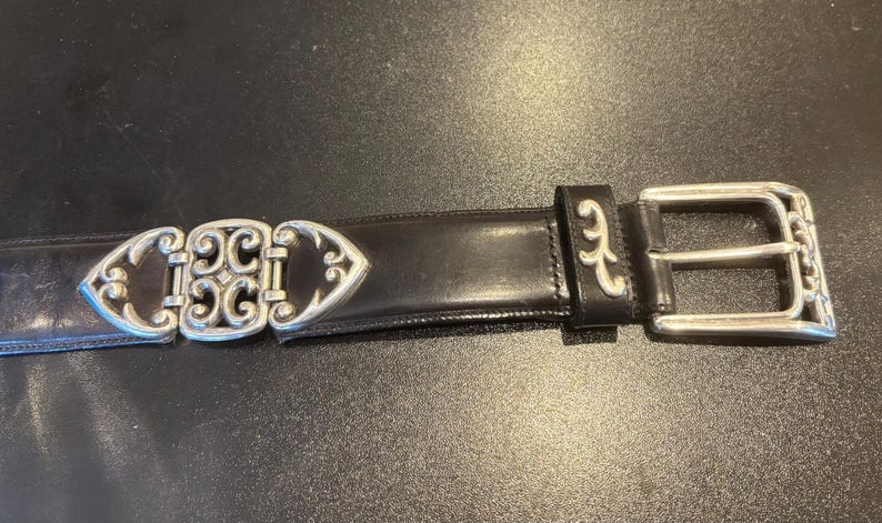 May include: Black leather belt with a silver-colored buckle and decorative accents. The belt features a detailed buckle design and a classic, elegant style. The belt is approximately 1.5 inches wide.