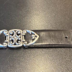 May include: A black leather belt with a decorative silver-colored buckle. The buckle features an ornate design with scrolling patterns. The belt has several holes for adjustment.