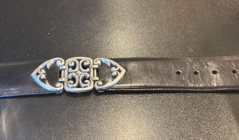 May include: A black leather belt with a decorative silver-colored buckle. The buckle features an ornate design with scrolling patterns. The belt has several holes for adjustment.