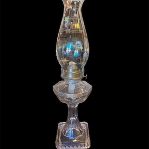 May include: A clear glass oil lamp featuring a square base with a Greek key pattern, a faceted pedestal, and a bulbous glass chimney. The lamp includes a silver-toned metal burner assembly with a visible white wick centered in the middle section.