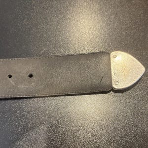 May include: A black leather belt with three holes and a silver-colored, triangular buckle. The belt is worn and shows signs of use. The buckle is attached with rivets.