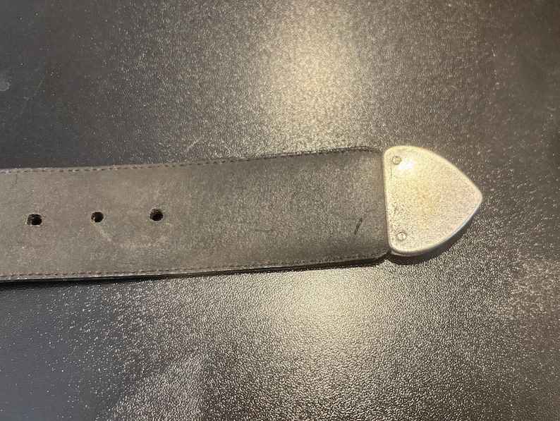 May include: A black leather belt with three holes and a silver-colored, triangular buckle. The belt is worn and shows signs of use. The buckle is attached with rivets.
