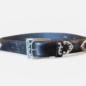 May include: Black leather belt with silver-toned buckle and decorative accents. The belt features a rectangular buckle with ornate detailing and decorative silver pieces along the belt's edge. A classic accessory.