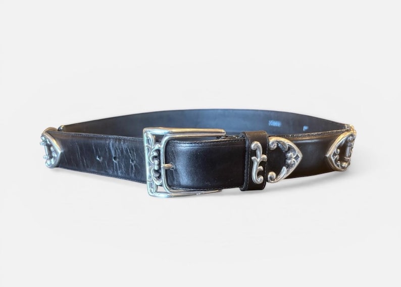 May include: Black leather belt with silver-toned buckle and decorative accents. The belt features a rectangular buckle with ornate detailing and decorative silver pieces along the belt's edge. A classic accessory.
