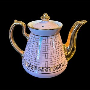 May include: A light pink porcelain teapot featuring a gold-colored basket weave pattern on the body, a gold-trimmed handle, a gold-colored spout, and a matching lid with a gold knob. A decorative band of small gold floral motifs encircles the base.