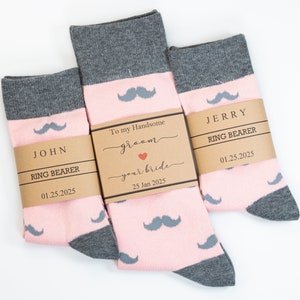 May include: Three pairs of socks with a mustache pattern in pink, gray, and white. Each pair is wrapped with a brown paper band that reads "JOHN RING BEARER 01.25.2025", "To my Handsome groom your bride 25 Jan 2025", and "JERRY RING BEARER 01.25.2025".