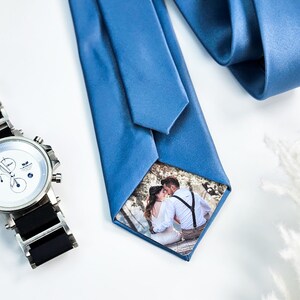May include: A blue silk tie with a small photo of a couple kissing inside the knot. The photo is surrounded by a white border. A silver watch with a black band is also in the image.