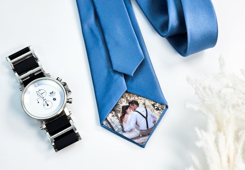 May include: A blue silk tie with a small photo of a couple kissing inside the knot. The photo is surrounded by a white border. A silver watch with a black band is also in the image.