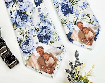 Puede incluir: Dos corbatas blancas con un patrón floral azul. Cada corbata tiene una foto de un hombre y un niño en un marco blanco.