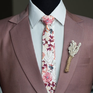 May include: A mauve suit jacket with a white collared shirt and a floral patterned tie. A small bouquet of dried white flowers is pinned to the lapel.