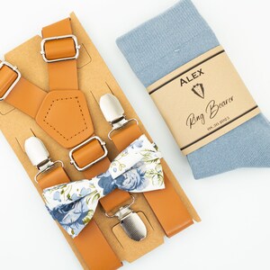 May include: A tan leather suspender set with a blue and white floral bow tie and a pair of light blue socks. The socks have a label that says "Alex Ring Bearer".