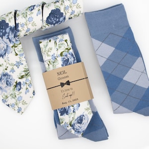 Könnte beinhalten: Ein Hochzeitsgeschenk-Set für den Bräutigam, bestehend aus einer blauen und weißen floralen Krawatte, einem passenden Einstecktuch und einem Paar blauen Argyle-Socken. Das Geschenk ist in einer braunen Papiertüte verpackt mit einem personalisierten Etikett, auf dem steht "Neil Groom It's time to Suit up!! Aug. 11, 2023".