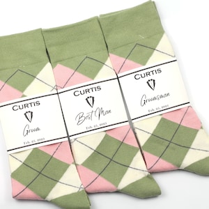 May include: Three pairs of socks with a green, pink, and white argyle pattern. Each pair has a white tag with black text that reads "CURTIS" and a title, "Groom", "Best Man", or "Groomsman", along with the date "Feb. 25, 2023".