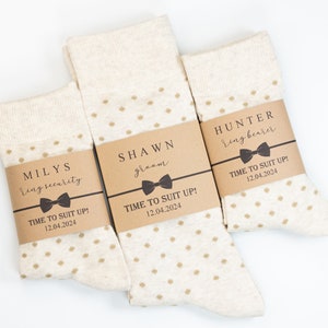 May include: Three pairs of white socks with brown polka dots and brown paper labels. The labels have the text "MILYS ring security", "SHAWN groom", and "HUNTER ring bearer" with a bow tie graphic and the text "TIME TO SUIT UP! 12.04.2024".