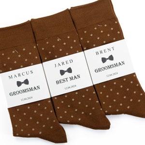 Peut inclure: Trois paires de chaussettes marrons à pois blancs. Chaque paire est accompagnée d'une étiquette blanche avec un nœud papillon noir et le texte "Best Man", "Groomsman" et "Groomsman" avec un nom et une date.