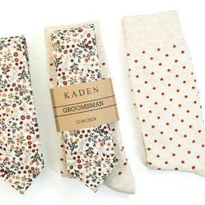 May include: A groomsman gift set featuring a beige tie and pocket square with a floral pattern in shades of brown and orange, and a pair of beige socks with small orange polka dots. The gift set is labelled with the name "Kaden" and the title "Groomsman" with the date "12.04.2024".