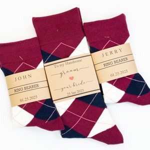 May include: Three pairs of red, white, and navy blue argyle socks with personalized tags. The tags are brown and have the text "To my Handsome groom your bride 25 Jan 2025", "JOHN RING BEARER 01.25.2025", and "JERRY RING BEARER 01.25.2025".