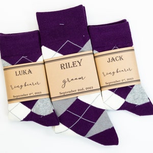 May include: Three pairs of socks with brown paper tags. The socks are purple, white, and gray argyle. The tags have the names Luka, Riley, and Jack, and the roles ring bearer and groom. The dates September 2nd, 2023 and September 2nd, 2023 are also on the tags.
