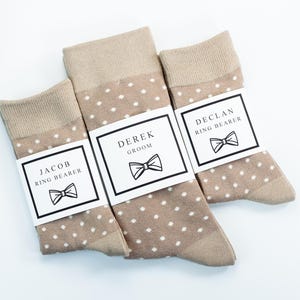 May include: Three pairs of brown socks with white polka dots. Each pair has a white label with black text and a bowtie graphic. The labels read "Derek Groom", "Jacob Ring Bearer", and "Declan Ring Bearer".