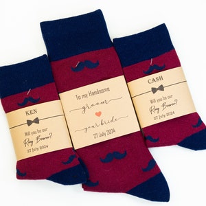 May include: Three pairs of socks with a mustache pattern. The socks are red and blue with a brown paper tag around each pair. The tags have text that says "Will you be our Ring Bearer?" and "To my Handsome Groom".