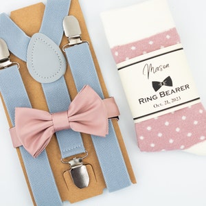 May include: A light blue suspender set with a matching pink bow tie and a pair of white and pink polka dot socks. The socks have a label that reads "Mason Ring Bearer Oct. 21, 2023".