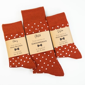May include: Three pairs of socks with white polka dots on a rust-colored background. Each pair has a brown tag with the text "Your service as" and the role of the wearer, such as "Ring Bearer" or "Groomsman", along with the name of the wearer and the date "Jan. 25, 2025".