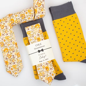 May include: A groomsman gift set featuring a yellow floral patterned tie, a yellow tie with a floral pattern, and yellow socks with grey polka dots. The gift tag reads "COLE Groomman It's time to Suit up!! Aug. 11, 2023"