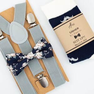 May include: A gray suspender set with a navy blue bow tie with a floral pattern. The set also includes a pair of white socks with a navy blue mustache pattern. The socks have a brown paper tag with the text "Alex Usher It's Time to Suit Up! March 9, 2024".