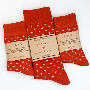 May include: Two pairs of orange socks with white polka dots. The socks have tags that say "Jerry Ring Bearer 01.25.2025" and "John Ring Bearer 01.25.2025". The larger sock has a tag that says "To my Handsome groom your bride 25 Jan 2025".