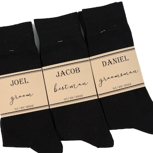 Może przedstawiać: Trzy pary czarnych skarpet z brązowymi papierowymi etykietami. Etykiety mają nazwy "Joel", "Jacob" i "Daniel" z tytułami "groom", "best man" i "groomsman". Data "05/29/2022" jest nadrukowana na każdej etykiecie.