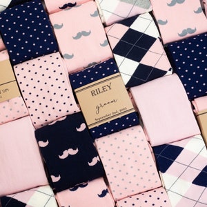 May include: A collection of pink and navy blue socks with different patterns, including polka dots, argyle, and moustaches. Each pair of socks has a brown paper tag with a name, the word "groom", and a date.