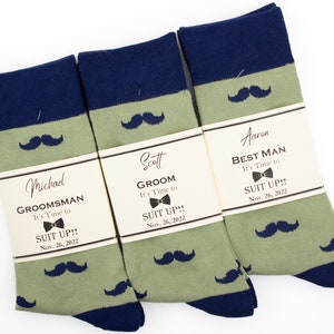 May include: Three pairs of green and navy blue socks with a mustache pattern. Each pair has a tag attached with the text "It's Time to Suit Up!" and the date "Nov. 26, 2022". The tags also include the names "Michad", "Scott", and "Aaron" and the titles "Groomsman", "Groom", and "Best Man".
