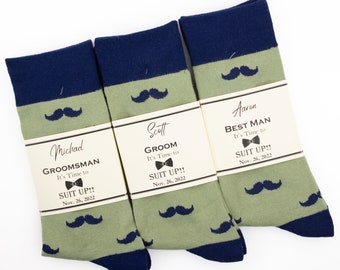 May include: Three pairs of green and navy blue socks with a moustache pattern. Each pair has a tag attached with the text "It's Time to Suit Up!" and the date "Nov. 26, 2022". The tags also include the names "Michad", "Scott", and "Aaron" and the titles "Groomsman", "Groom", and "Best Man".