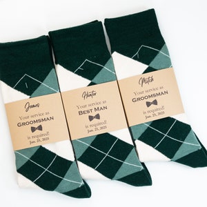 May include: Three pairs of green and white argyle socks with brown paper tags attached. The tags have the names "James", "Hunter", and "Mitch" printed on them, along with the text "Your service as GROOMSMAN is required! Jan. 25, 2025".