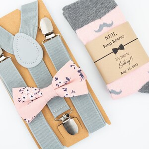 May include: A gray suspender set with a pink bow tie with white floral print and a pair of pink and gray socks with a mustache design. The socks have a brown paper tag with the text "NEIL Ring Bearer It's time to Suit up!! Aug. 12, 2023"
