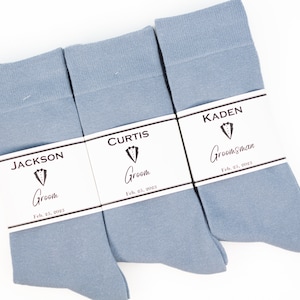 May include: Three pairs of light blue socks with white labels. The labels have the names "Jackson", "Curtis", and "Kaden" with the words "Groom" and "Groomsman" and the date "Feb. 25, 2023".