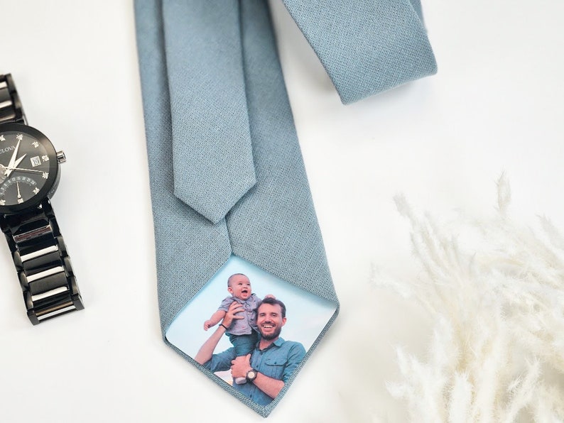 Puede incluir: Una corbata azul claro con un tejido texturizado. La parte inferior de la corbata presenta una fotograf&iacute;a en color de un hombre sosteniendo a un beb&eacute;. Un reloj de pulsera negro con una correa negra es visible en el fondo.