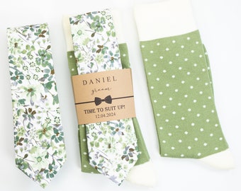 May include: A green and white floral patterned tie and a pair of green socks with white polka dots. The socks and tie are packaged together with a brown tag that reads "DANIEL groom TIME TO SUIT UP! 12.04.2024".
