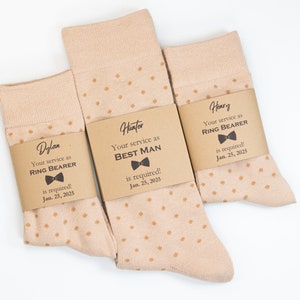 Puede incluir: Tres pares de calcetines con lunares marrones y etiquetas personalizadas. Las etiquetas son marrones con texto blanco y una pajarita negra. El texto en las etiquetas dice "Su servicio como padrino de boda es requerido! Jan. 25, 2025", "Su servicio como portador de anillos es requerido! Jan. 25, 2025" y "Su servicio como portador de anillos es requerido! Jan. 25, 2025".