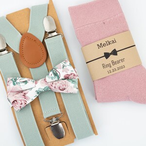 May include: A light green suspender set with a floral bow tie and a pair of pink socks. The socks have a brown label that says "Melkai Ring Bearer 12.23.2023".