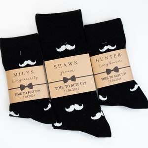 May include: Three pairs of black socks with white mustache designs and brown paper tags. The tags have the text "MILYS ring security", "SHAWN groom", and "HUNTER ring bearer" with the date "12.04.2024" and the phrase "TIME TO SUIT UP!" on each.