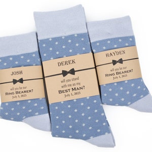 May include: Three pairs of blue socks with white polka dots and brown paper tags with text that reads "JOSH will you be our RING BEARER? July 1, 2023", "DEREK will you stand with me as my BEST MAN? July 1, 2023", and "HAYDEN will you be our RING BEARER? July 1, 2023".
