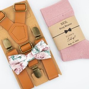 Op de afbeelding: Een bruine leren bretelsset met een bloemenstrik en een paar roze sokken met een bruin label waarop staat "NEIL Ring Bearer It's time to Suit up!! Aug. 12, 2023"