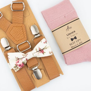 Op de afbeelding: Een bruine leren bretelsset met een bloemenstrik en een paar roze sokken. De sokken hebben een bruin papieren etiket met de tekst "Alex Usher It's Time to Suit Up!! March 9, 2024".