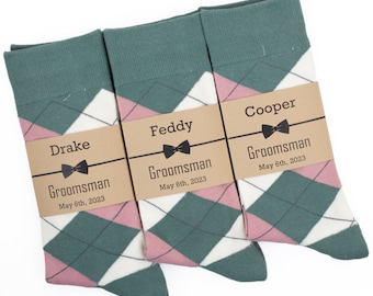 May include: Three pairs of argyle socks with pink, white and green diamond patterns. Each pair has a brown tag with the text "Groomsman" and a name, "Drake", "Feddy", or "Cooper", and the date "May 6th, 2023".