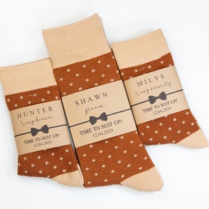 Puede incluir: Tres pares de calcetines con lunares marrones y puños y dedos de los pies beiges. Cada par tiene una banda de papel marrón con el texto: "HUNTER ring bearer", "SHAWN groom" y "MILYS ring security". También está impreso el texto "TIME TO SUIT UP! 12.04.2024".