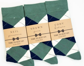 May include: Three pairs of green, white and navy blue argyle socks with brown paper tags. The tags have the text "TIME TO SUIT UP!" and the date "12.04.2024". The tags also have the names "NEIL groomsman", "SHAWN groom", and "CODY best man".