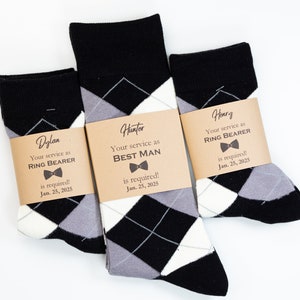May include: Three pairs of black and white argyle socks with brown paper tags. The tags have the names Dylan, Hunter, and Henry, and the text "Your service as Ring Bearer is required! Jan. 25, 2025" or "Your service as Best Man is required! Jan. 25, 2025".