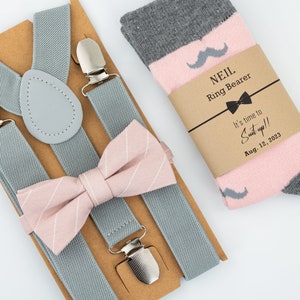 May include: Gray suspenders with a pink and white striped bow tie and a gray and pink sock with a mustache design. The sock has a brown paper tag that says "Neil Ring Bearer It's time to Suit up!! Aug. 12, 2023"