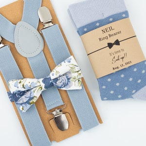 May include: A light blue suspender set with a floral bow tie and a pair of light blue socks with white polka dots. The socks have a brown paper tag with the text "NEIL Ring Bearer It's time to Suit up!! Aug. 12, 2023"